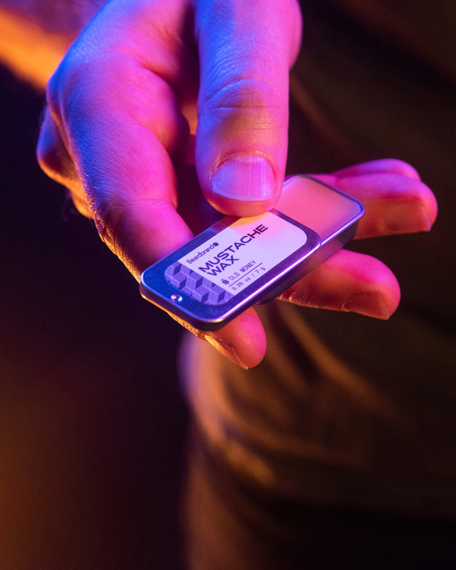 A tin of Beardbrand Mustache Wax being slid open by a person's hand highlighted in vibrant lighting.