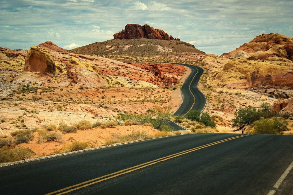 A winding desert highway curves through vibrant red and yellow sandstone formations, with rugged mesas rising in the background under a cloudy blue sky.