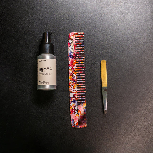 Beard grooming set featuring Beardbrand Old Money Beard Oil, a colorful marbled large comb, and metal tweezers with a gold handle on a dark background.