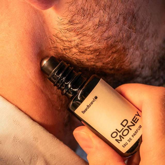 Close-up of stubbled lower face with Beardbrand Old Money Eau de Parfum roll-on bottle held against the skin.