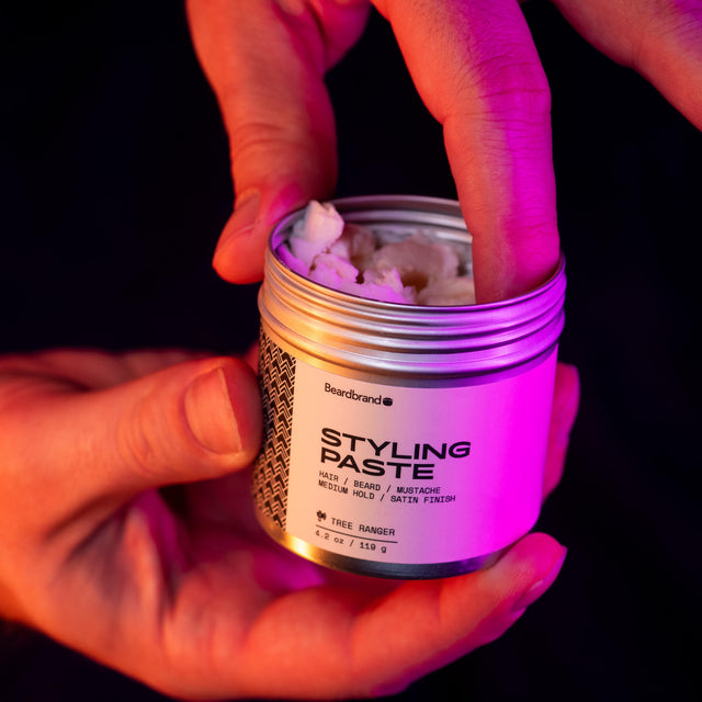 A hand holding an open jar of Beardbrand Styling Paste illuminated by pink neon light, against a dark background.