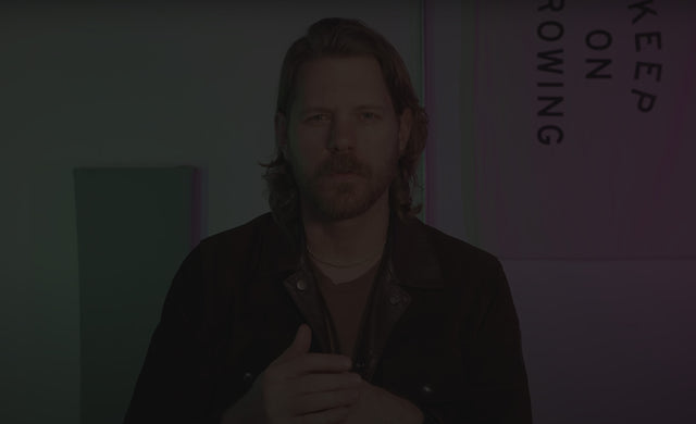 Eric Bandholz in a video still standing behind the camera with his hands together in mid conversation. He's standing in front of a vertically hung "Keep on Growing" white flag with the text in black. The wall behind him is lit up in green and purple vibrant but filtered hues.