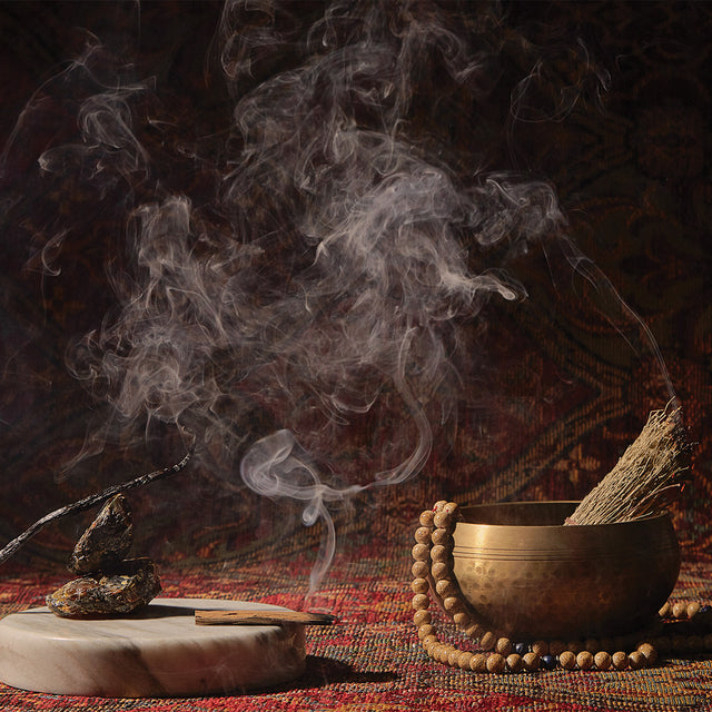 A still life of items reminiscent of Temple Smoke’s fragrance notes: a stick of palo santo burning with the smoke swirling across a scene of a bowl and beads, set up on a tapestry.