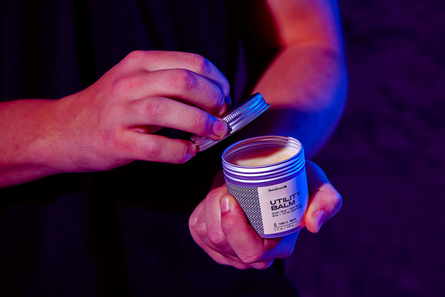 A person holds an open container of Beardbrand Utility Balm with the lid in their other hand.