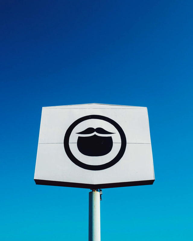 The Beardbrand Barbershop sign with a black circle beard icon on a white stretched hexagon in front of a cloudless electric blue sky in Austin, TX.