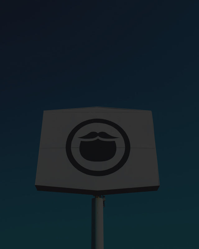 Beardbrand Barbershop signage is a white hexagon shape stretched to nearly be a square and has the Beardbrand circle beard icon on the front in black. The sign is on top of a metal post and is in front of a cloudless electric blue sky in Austin, Texas. The sky is a bright gradient of light blues from the bottom to darker hues of blue near the top left corner of the image.