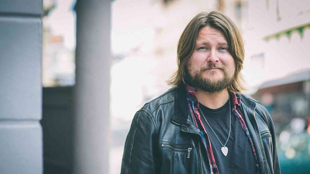 Man with Rock n Roll style standing outside. He has shoulder-length brown hair and is searing a leather jacket. 