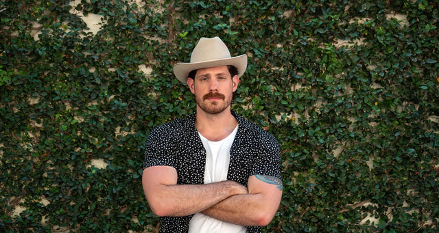 Man wearing a Stetson Open Raod is standing with arms crossed in front of an ivy-covered wall.