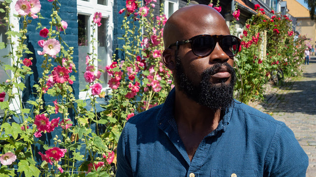 Manscaped Black man with a bald head and thick beard is wearing dark sunglasses and standing in front of flowers. 