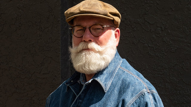 A bespectacled older gentleman with a magnificently shaped white beard and a handlebar mustache, and wearing a denim jacket. 