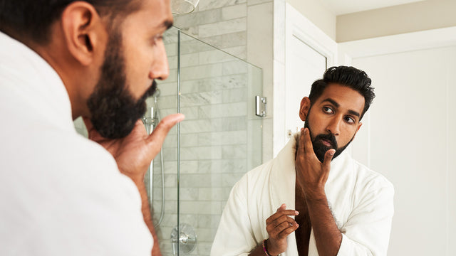 Man with a brown beard is standing in the bathroom and looking in the mirror. 