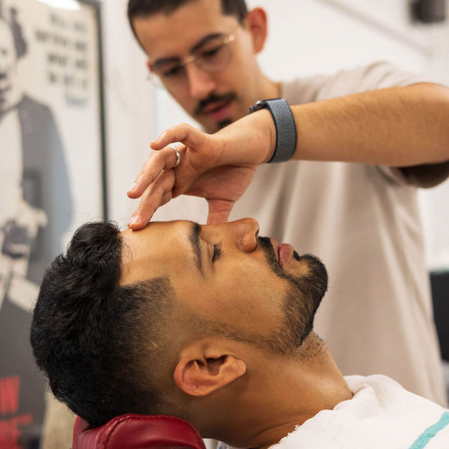 A barber precisely lining up a client’s hairline while the client reclines in a vintage red barber chair.