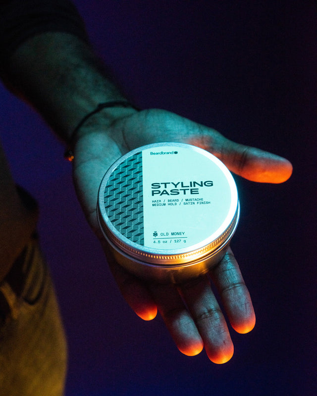 A canister of Beardbrand Styling Paste held in a person's hands with vibrant lighting so the top lid label is showing.