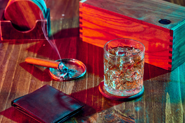 Whiskey in a crystal glass with ice on wood; smoking cigar nearby, wooden box in back, leather wallet in forefront.