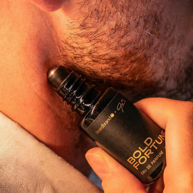 Close-up of stubbled lower face with Beardbrand Bold Fortune Eau de Parfum roll-on bottle held against the skin.