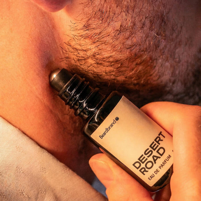 Close-up of stubbled lower face with Beardbrand Desert Road Eau de Parfum roll-on bottle held against the skin.