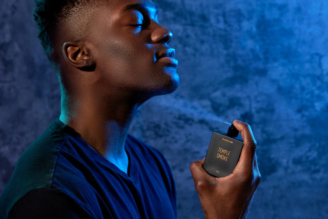 A man under blue lighting in front of a textured backdrop spraying the scent of Temple Smoke Eau de Parfum under his chin and enjoying the scent.