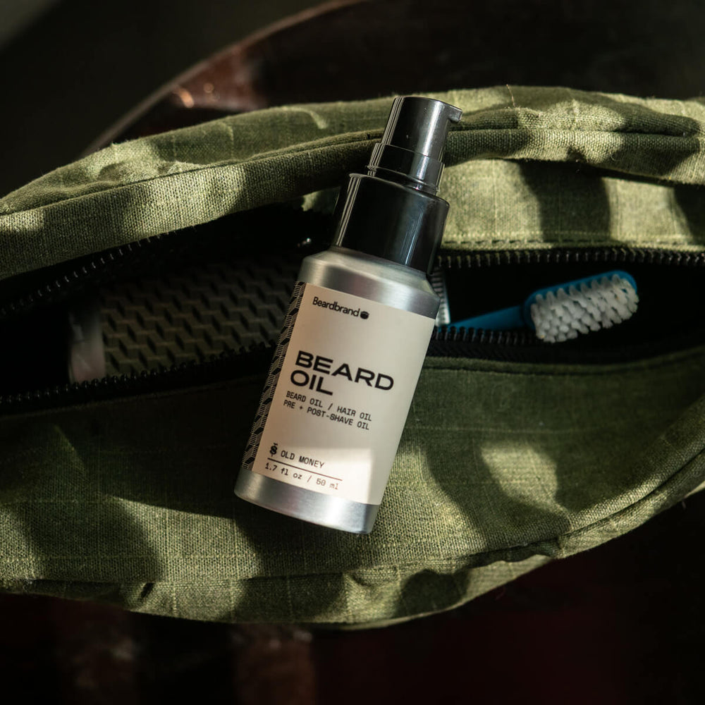 Beardbrand Old Money Beard Oil with pump dispenser on green canvas dopp bag in dappled sunlight.