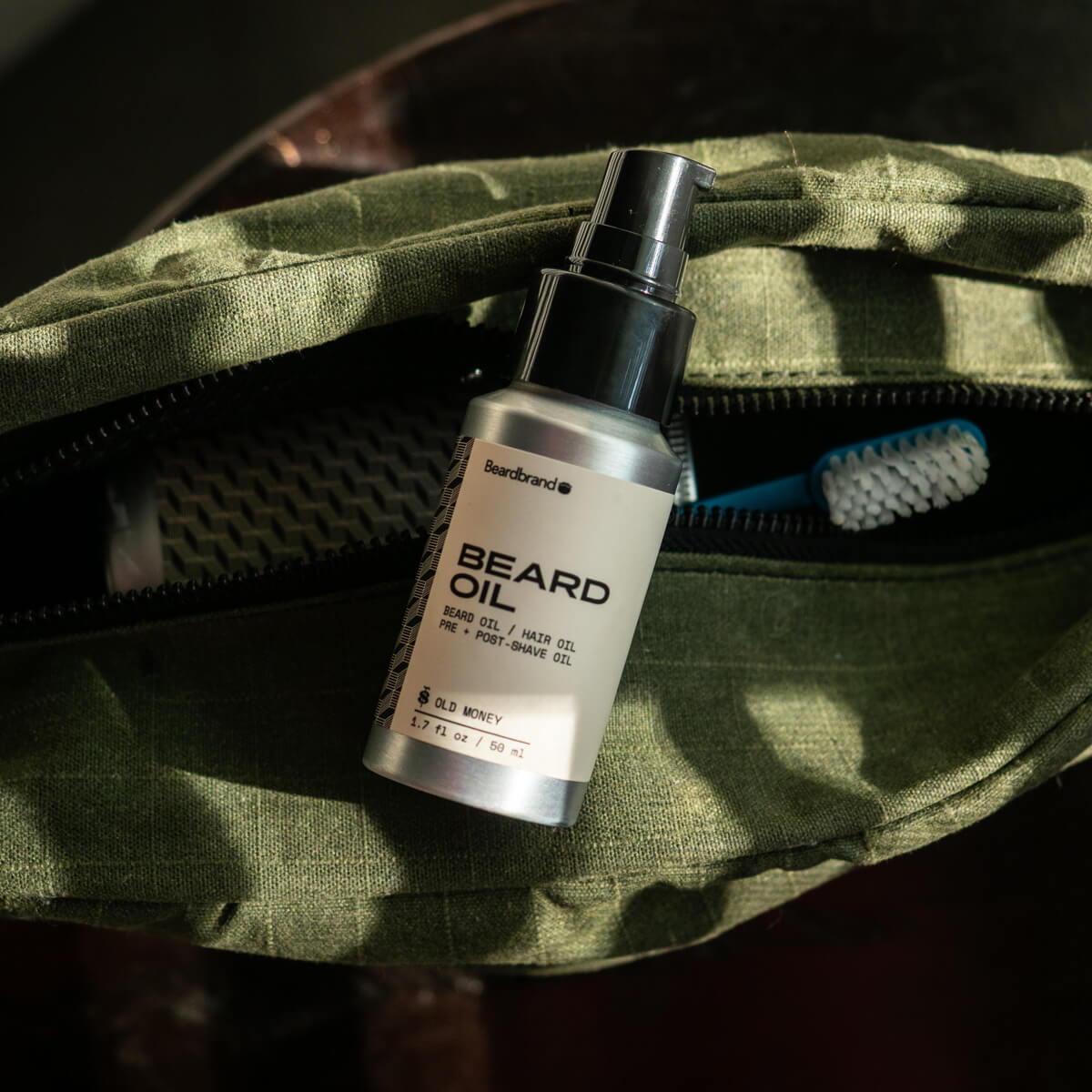 Beardbrand Old Money Beard Oil with pump dispenser on green canvas dopp bag in dappled sunlight.
