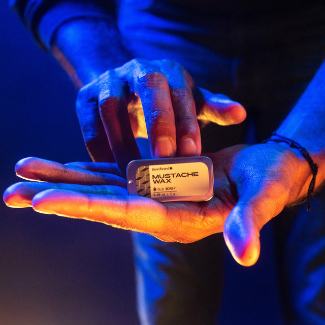 A tin of Beardbrand Mustache Wax being displayed in a person's hand highlighted in vibrant lighting.
