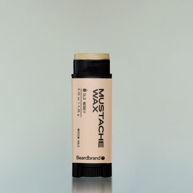 An Old Money Mustache Wax in a black plastic tube with the cap off showing the wax against a grey background.