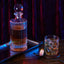 An arrangement of items on a wooden table in moody lighting: a crystal decanter on a metal tray, a glass of whiskey with ice, and a pocket watch.