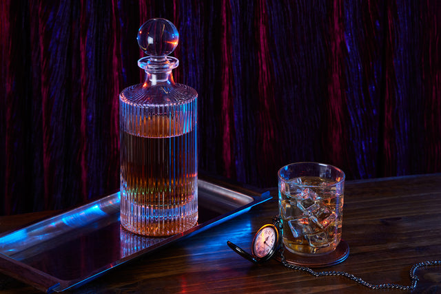 An arrangement of items on a wooden table in moody lighting: a crystal decanter on a metal tray, a glass of whiskey with ice, and a pocket watch.