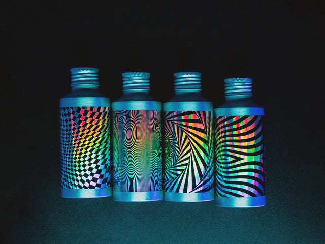 Four aluminum Beardbrand Utility Oil bottles in a untidy row on a dark surface, with holographic labels and black psychedelic designs in dim lighting.