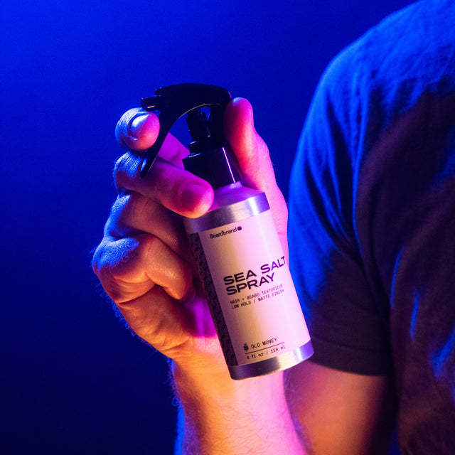 A person holding Beardbrand Sea Salt Spray, showing the front label, in vibrant lighting.