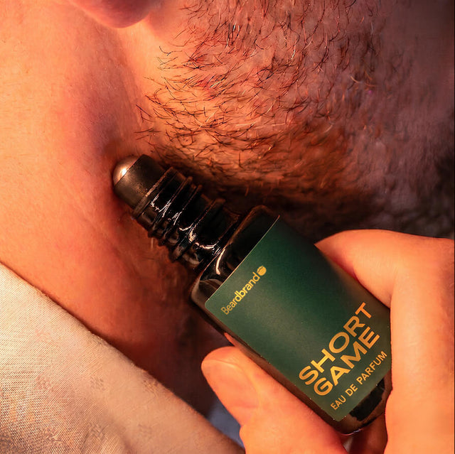 Close-up of stubbled lower face with Beardbrand Short Game Eau de Parfum roll-on bottle held against the skin.