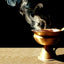 Golden brass incense burner with wisps of white and blue smoke rising against a black background. The ornamental vessel sits on a light beige surface, casting a soft shadow.