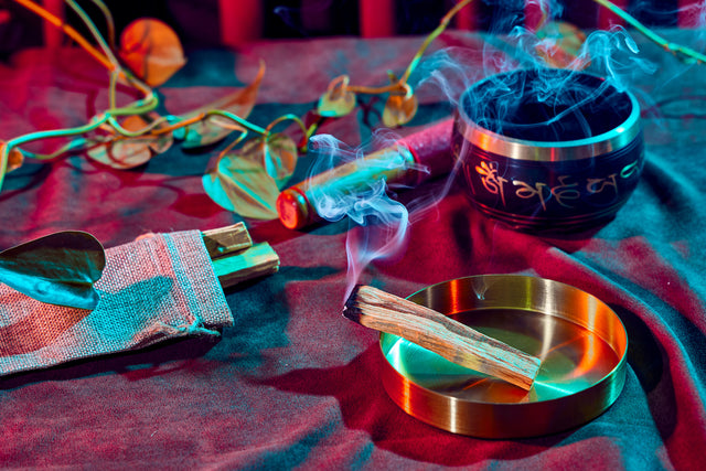 Smoky incense on metal plate; singing bowl, plant vines, set on top of woven fabric against red-blue backdrop.