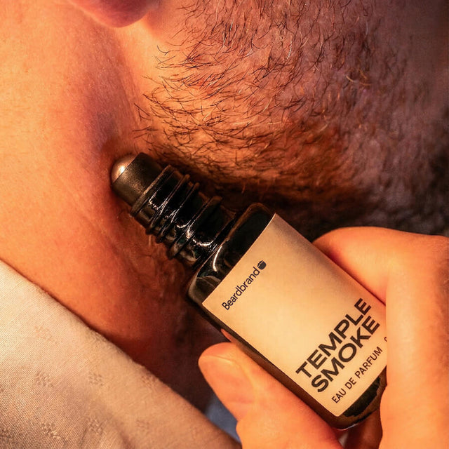 Close-up of stubbled lower face with Beardbrand Temple Smoke Eau de Parfum roll-on bottle held against the skin.