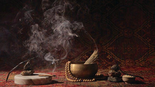 A still life of items reminiscent of Temple Smoke’s fragrance notes: a stick of palo santo burning with the smoke swirling across a scene of a bowl and beads, set up on a tapestry.