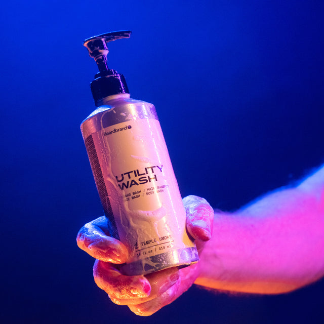 A person holding a sudsy container of Beardbrand Utility Wash in one hand, showing the front of the label, in vibrant lighting.