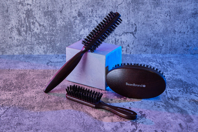 Three Beardbrand grooming brushes in different styles arranged on a textured surface with blue lighting.