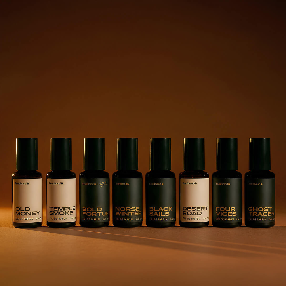 Beardbrand eau de parfum cologne lineup featuring Old Money, Temple Smoke, Bold Fortune, Norse Winter, Black Sails, Desert Road, Four Vices and Ghost Tracer bottles on a warm amber background.