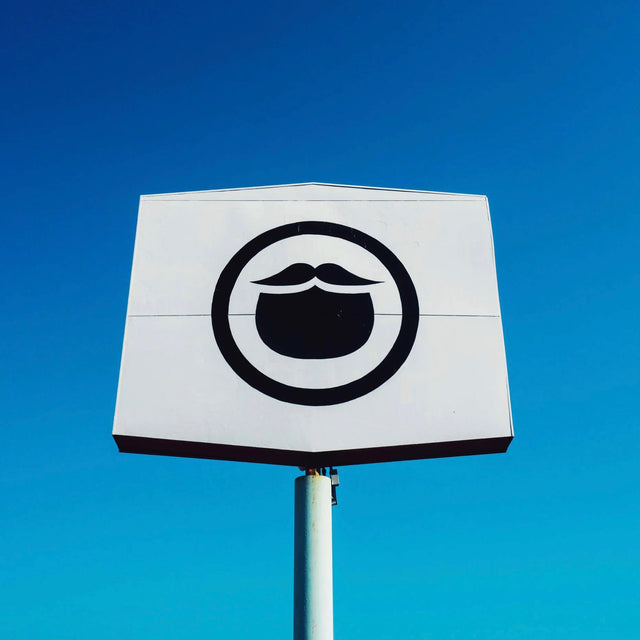 The Beardbrand Barbershop sign with a black circle beard icon on a white stretched hexagon in front of a cloudless electric blue sky in Austin, TX.