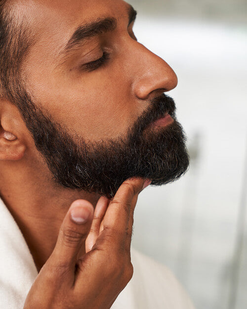 Close-up of a man with a well-groomed, thick black beard touching his chin while inspecting his facial hair. He is wearing a white robe, and the background is softly blurred.