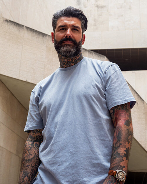 A bearded man with salt-and-pepper hair and a full beard stands confidently in an outdoor urban setting. He is wearing a light blue t-shirt and has tattooed arms. A watch is visible on his wrist.